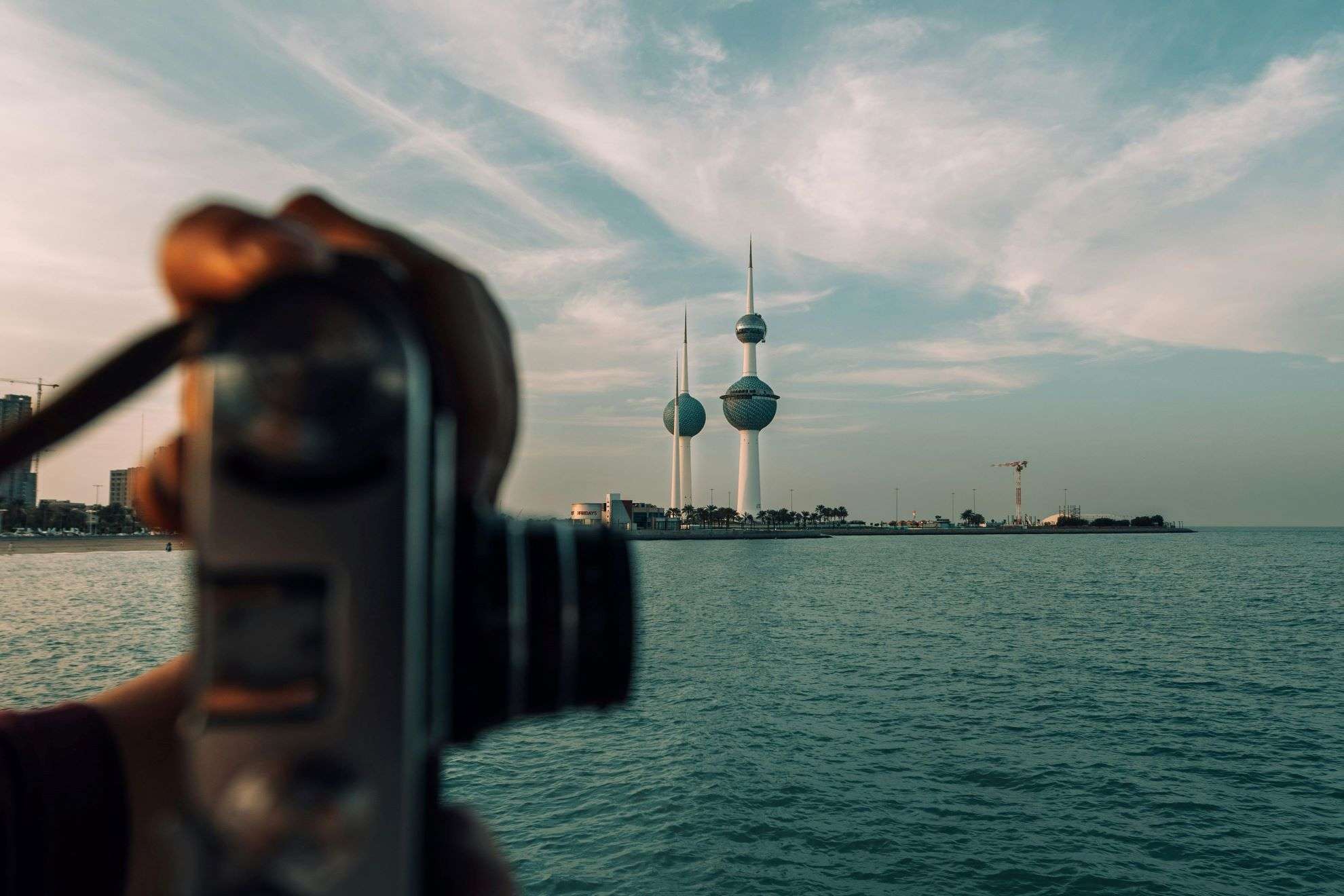 A person holds a vintage camera, capturing the beauty of the Kuwait Towers against a picturesque waterfront. Turath